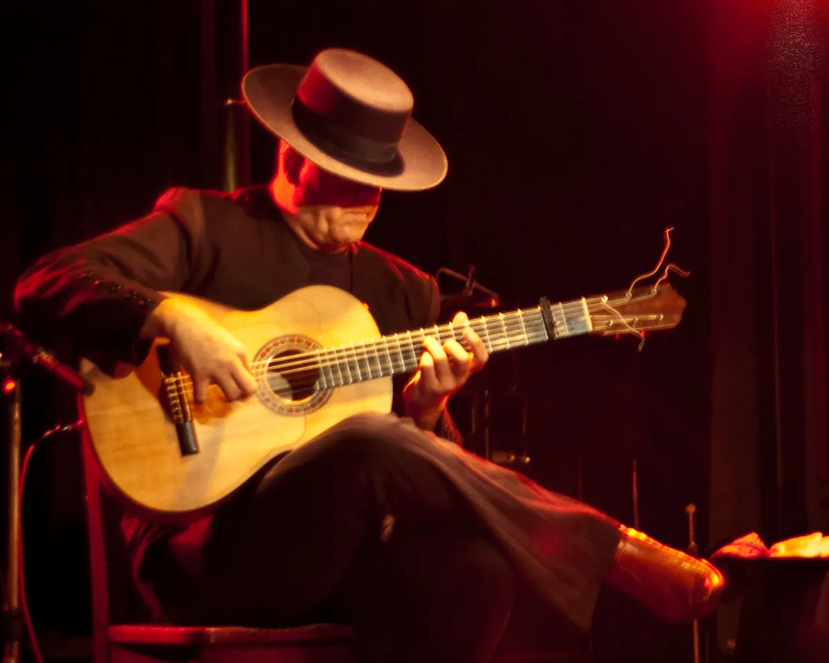 A flamenco guitarist in a wide-brimmed hat playing a blonde guitar in red stage light